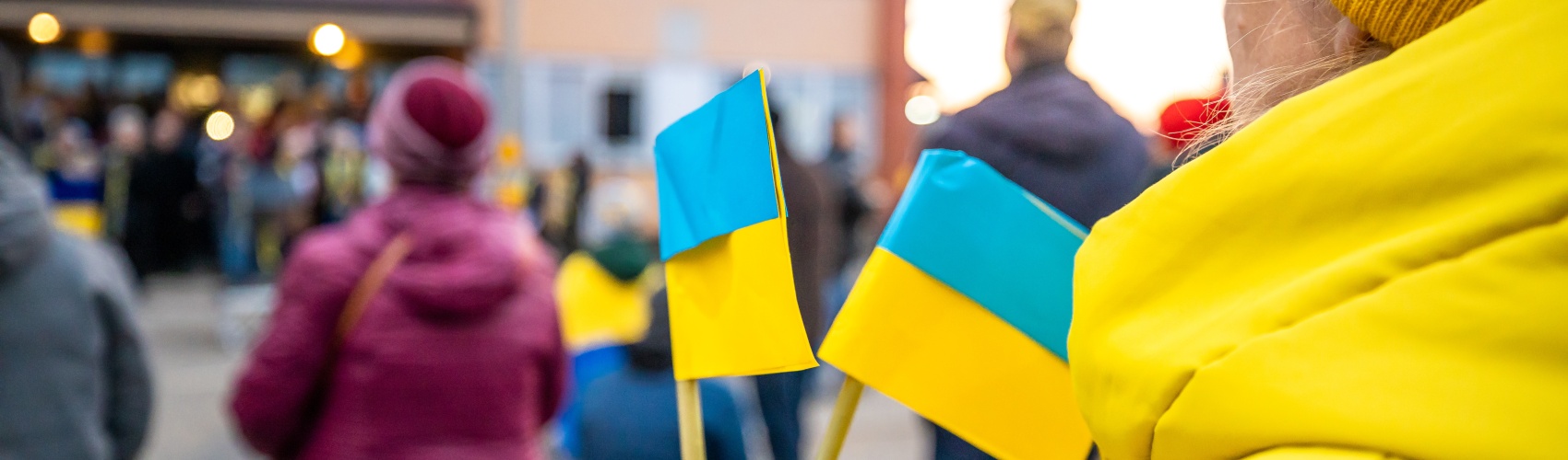  A woman in a bright yellow coat and mustard hat holds two small Ukrainian flags. In the background there are other protestors too. 