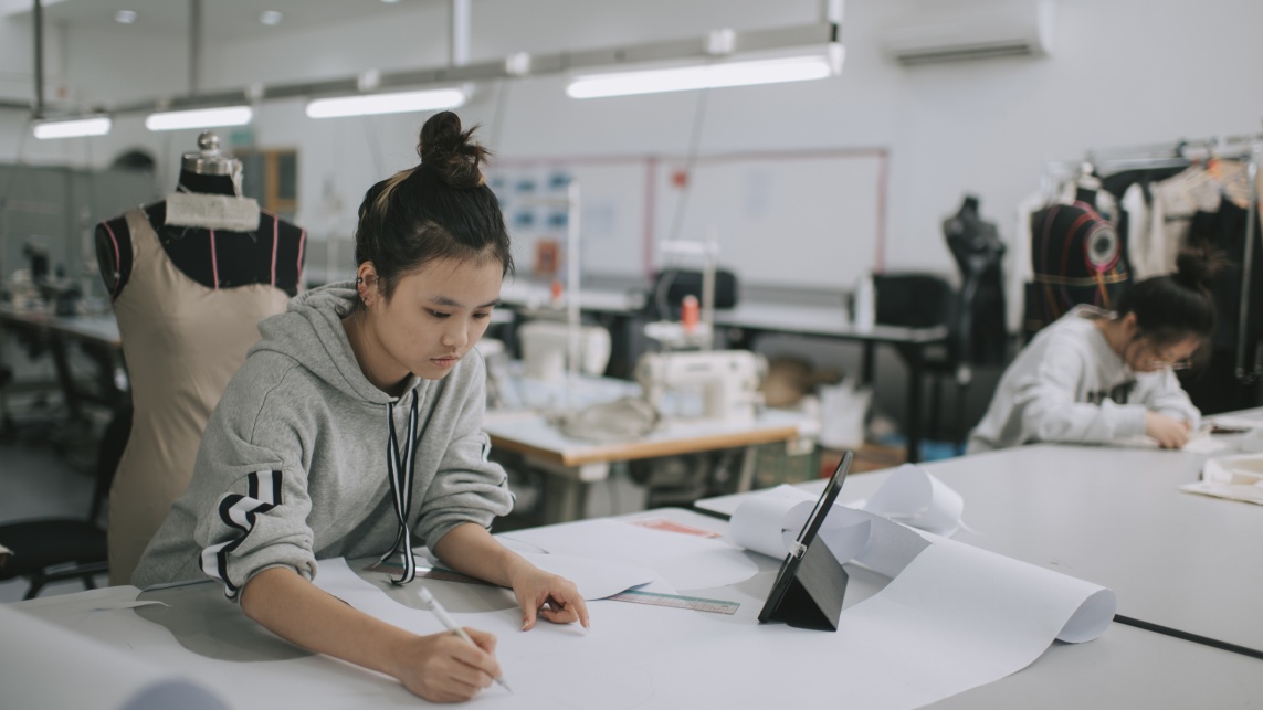 asian chinese female fashion college student writing down measurement and drawing sewing pattern doing clothing project at college workshop