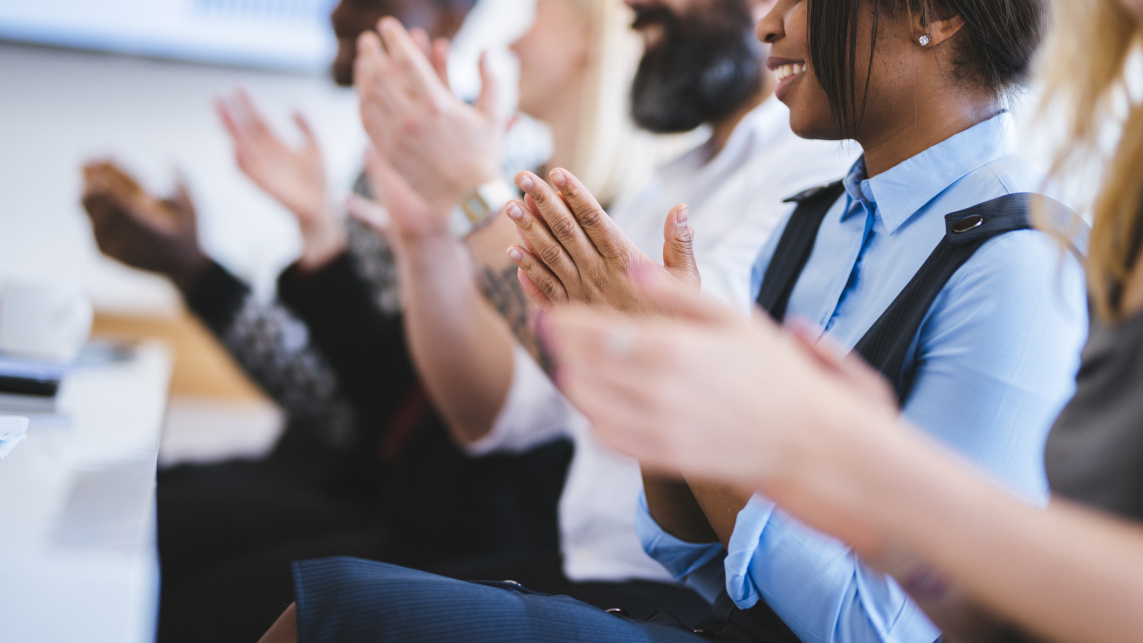 People sitting watching a presentation clapping their hands. They sit in front of a white desk.