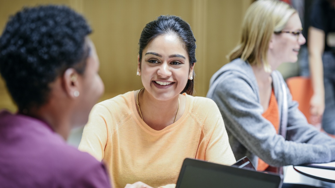 Cheerful young woman listening to friend in college classroom, smiling