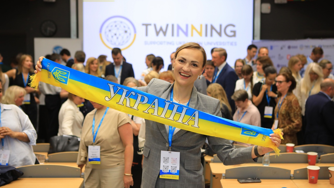 A woman poses with a scarf showing the colours of the Ukraine flag and text stating 'Ukraine' written in Ukrainian at the Second International Twinning Conference.