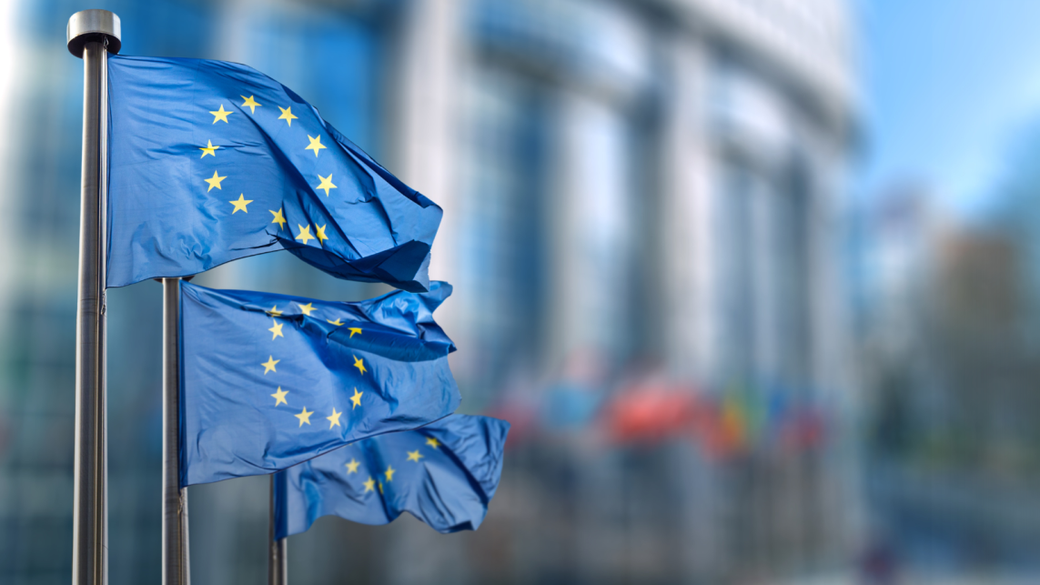 Three European Union flags flying at full mast against the blurred backdrop of a building.