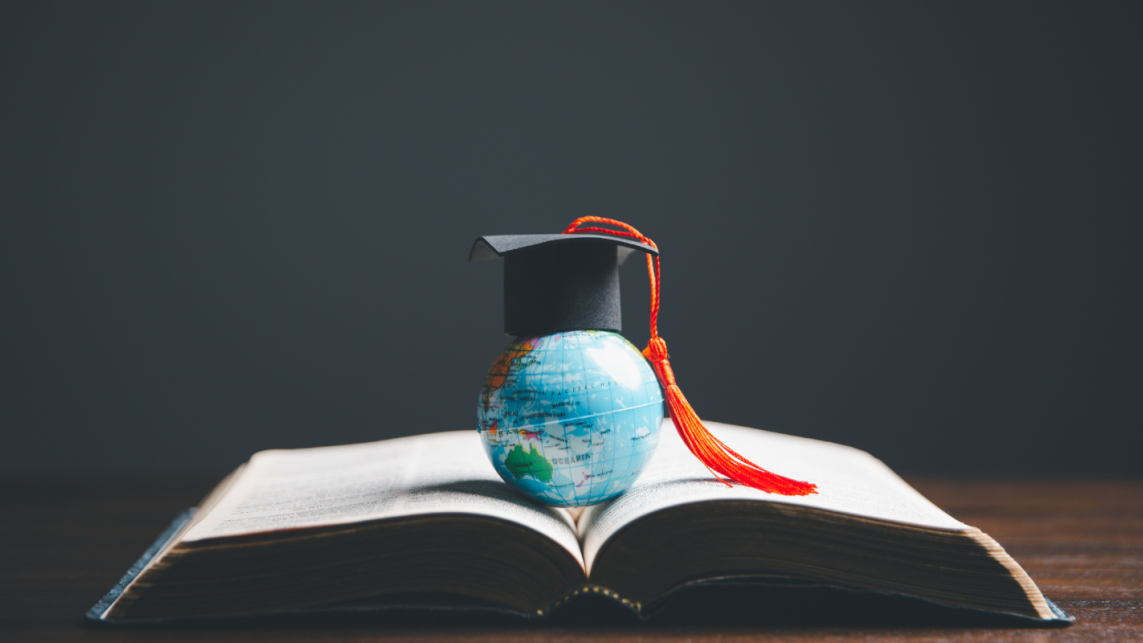 A book lies on a table, open, with a small globe and a graduation cap resting on top of it.