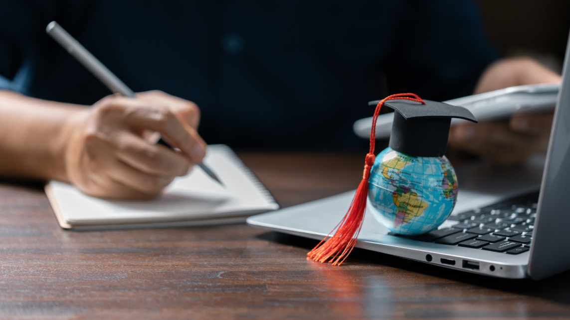 A small globe rests on a laptop keyboard with a graduation cap on top. To the left, a hand can be seen making notes in a notebook.