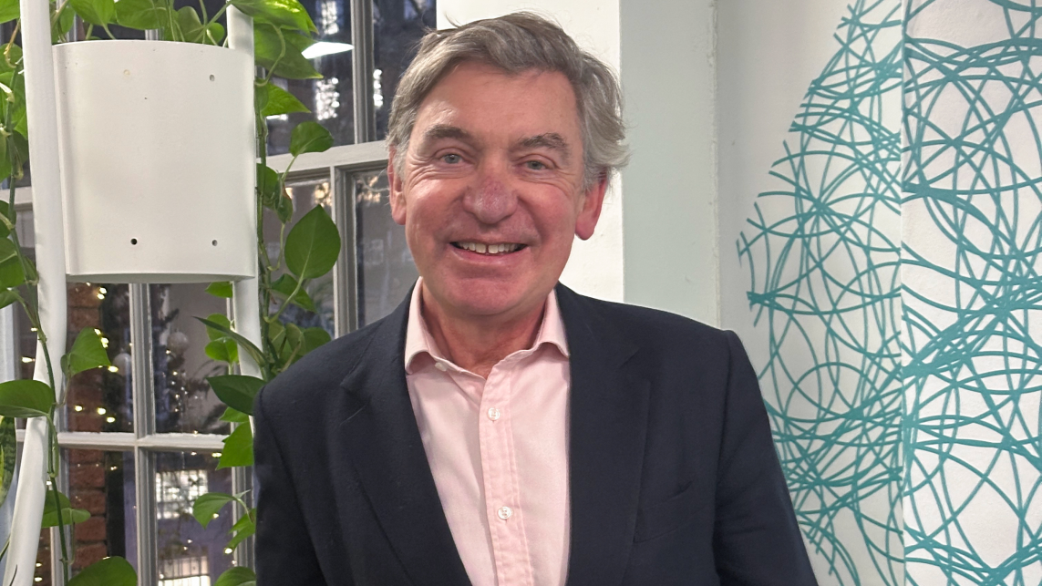 Sir Nigel Carrington standing in front of a white background with plants. Nigel wears a black suit and salmon shirt.