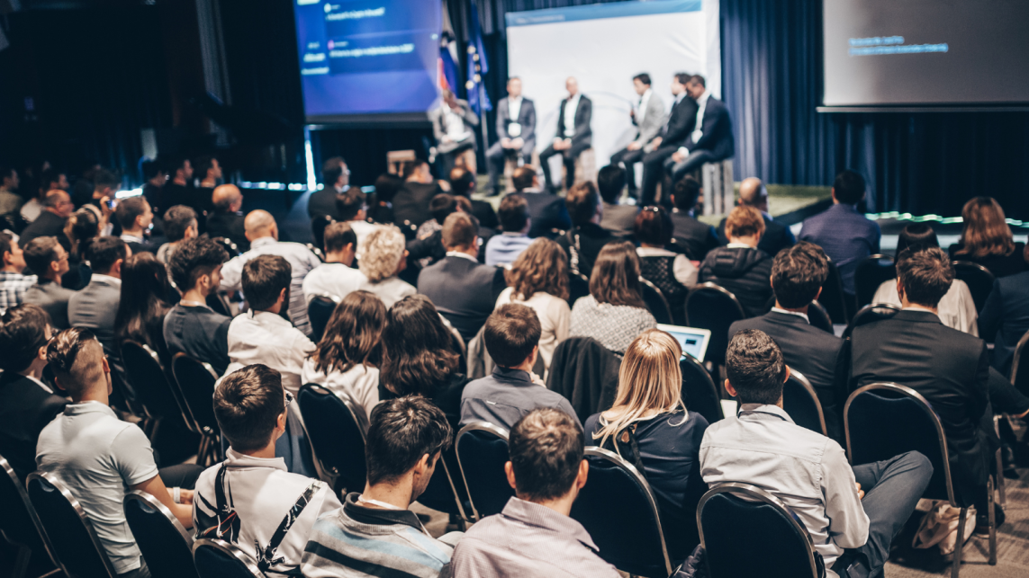 A conference room full of attendees, focusing on the stag where there is a panel of 5 speakers