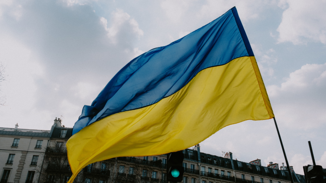 Ukraine flag blowing in the wind against a backdrop of sky and buildings.