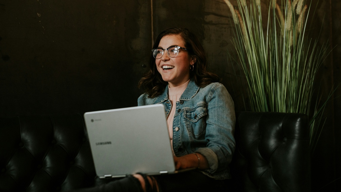 A student with a laptop sits on a sofa smiling 