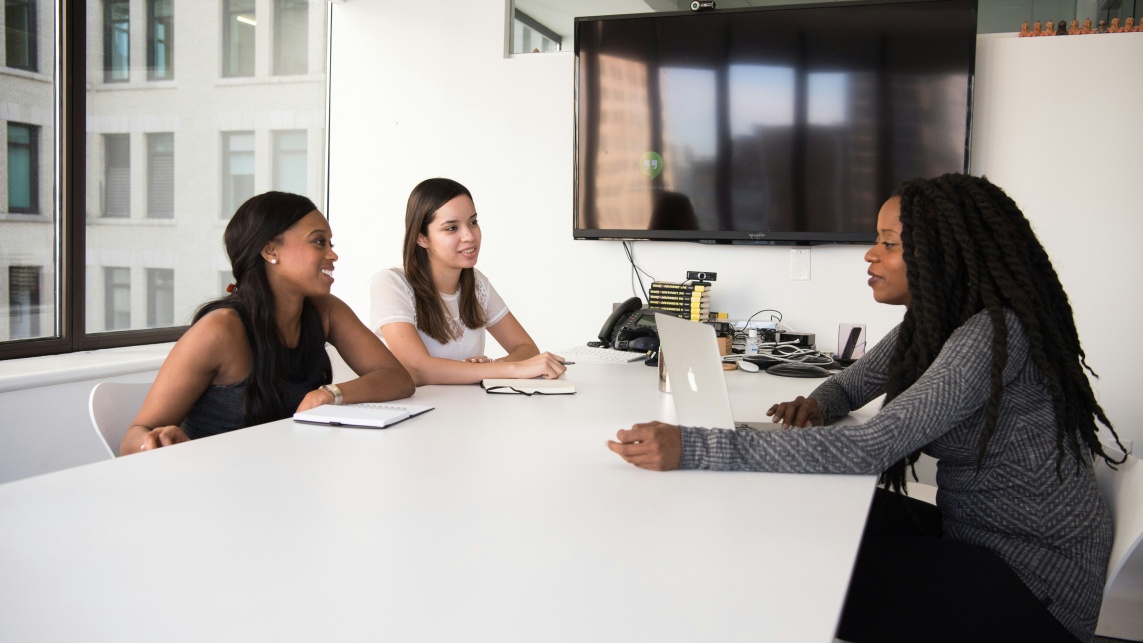 Three people sit at a desk in an office discussing someting, with laptops and notepads in front of them 