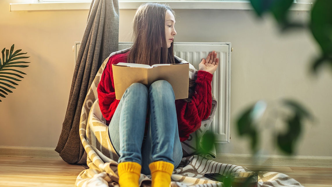 Female sits in front of a radiator, wrapped in a blanket, reading a book