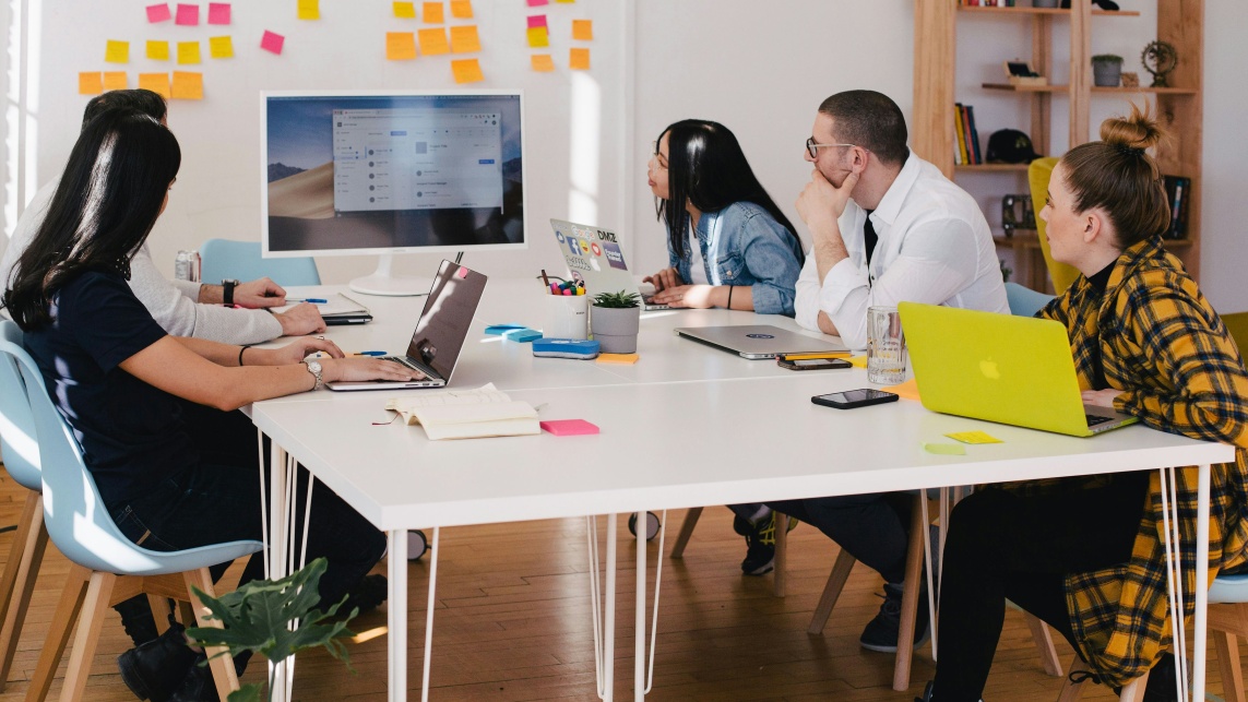 Four people sit in a room either side of a table, looking at a computer screen. Post-it notes are on the wall behind it. 