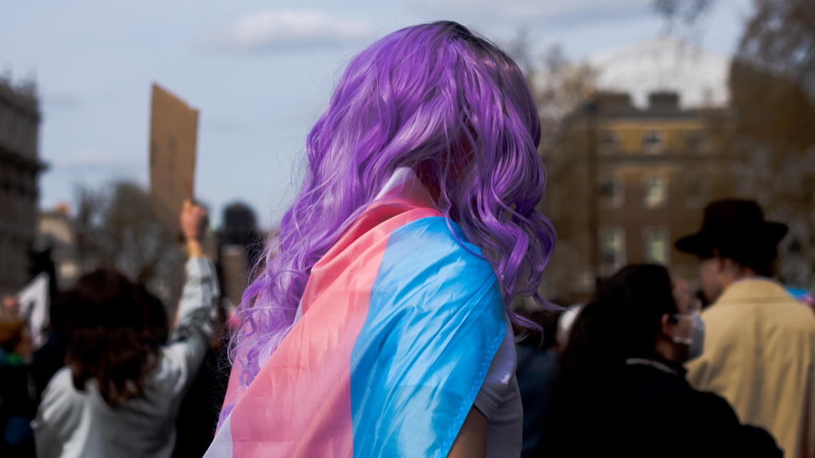 Person with purple hair and trans pride flag around their shoulders at a protest