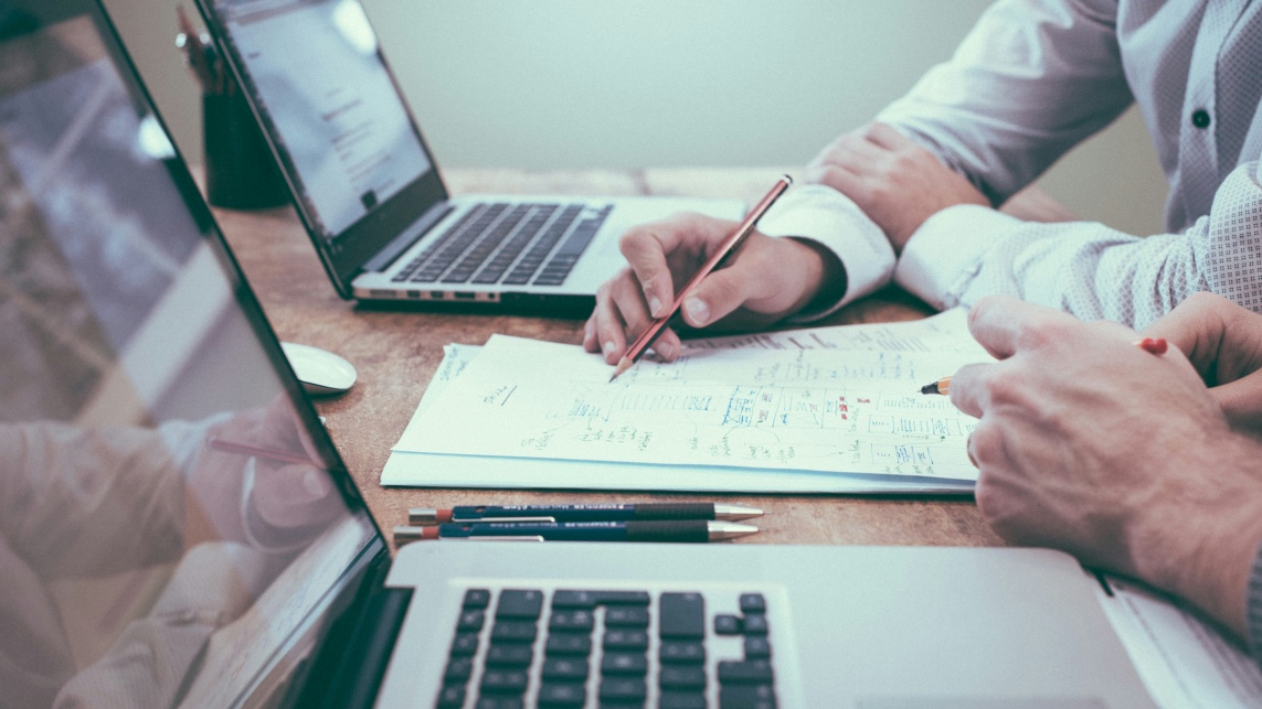 A person holding a pencil over some paper next to a colleague with two laptops, one for each person