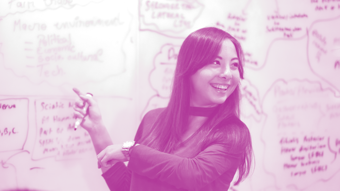 A young woman is gesturing to notes written on a whiteboard and smiling