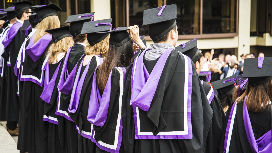 Back view of a university graduation ceremony. The graduates wear black and purple gowns and graduation caps.