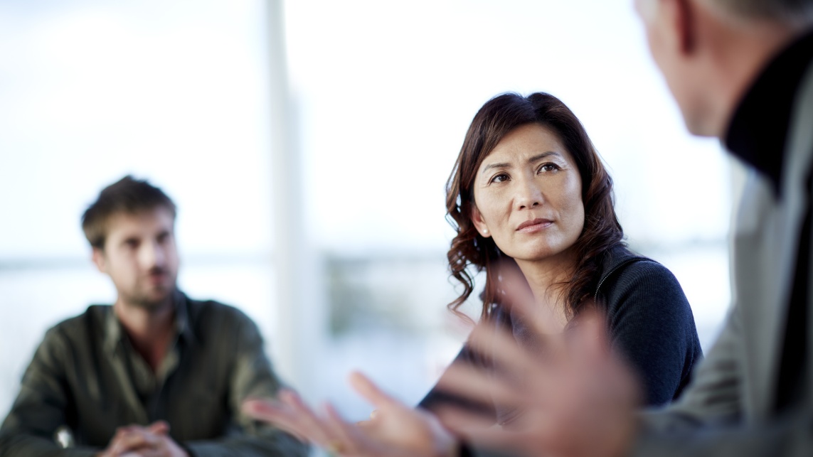 Three people in an office sit at a desk in conversation. A woman in the middle looks at the man speaking to the right who is blurred.