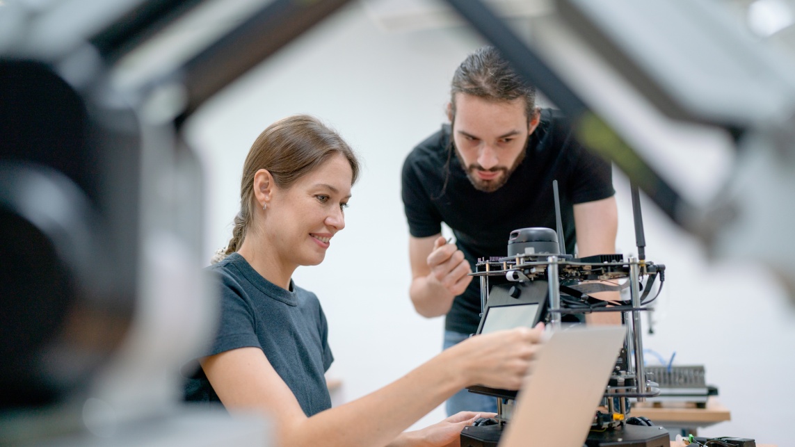 Two students at a desk with engineering equipment in front of them. The female student is adjusting the equipment and the male student leans over to observe.