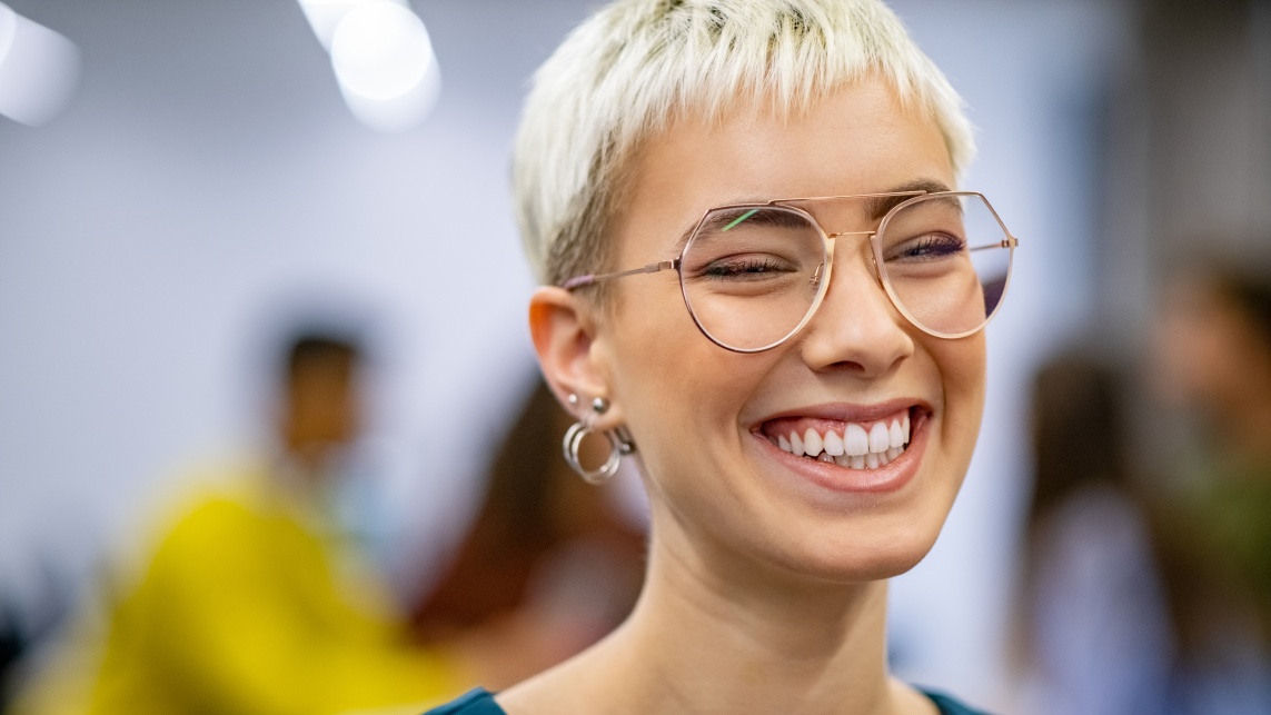 A woman smiles with her eyes shut. She has short blonde hair, wears large round glasses and a blue top.