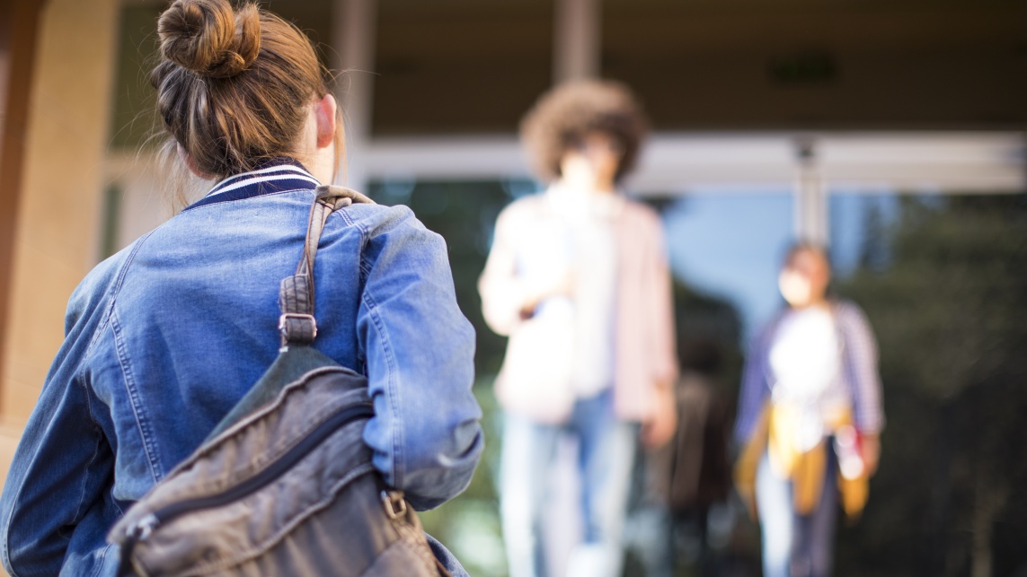 Student walks into campus building holding a background. She wears a denim jacket and stripped shirt.
