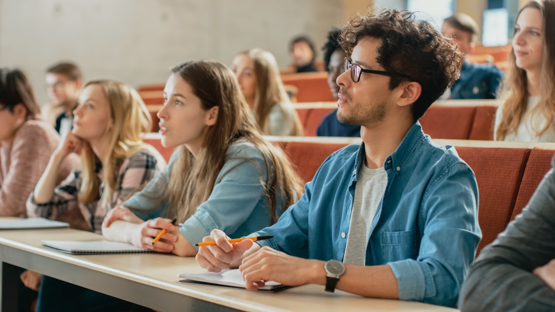 Students in a lecture hall looking towards the front of the room. Students in the front row hold pens with notepads in front of them.