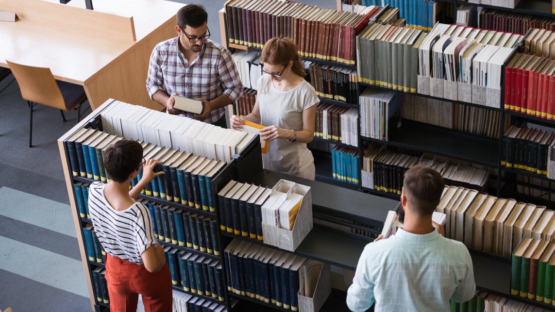 Four students in a library, browsing books from a bookcase.