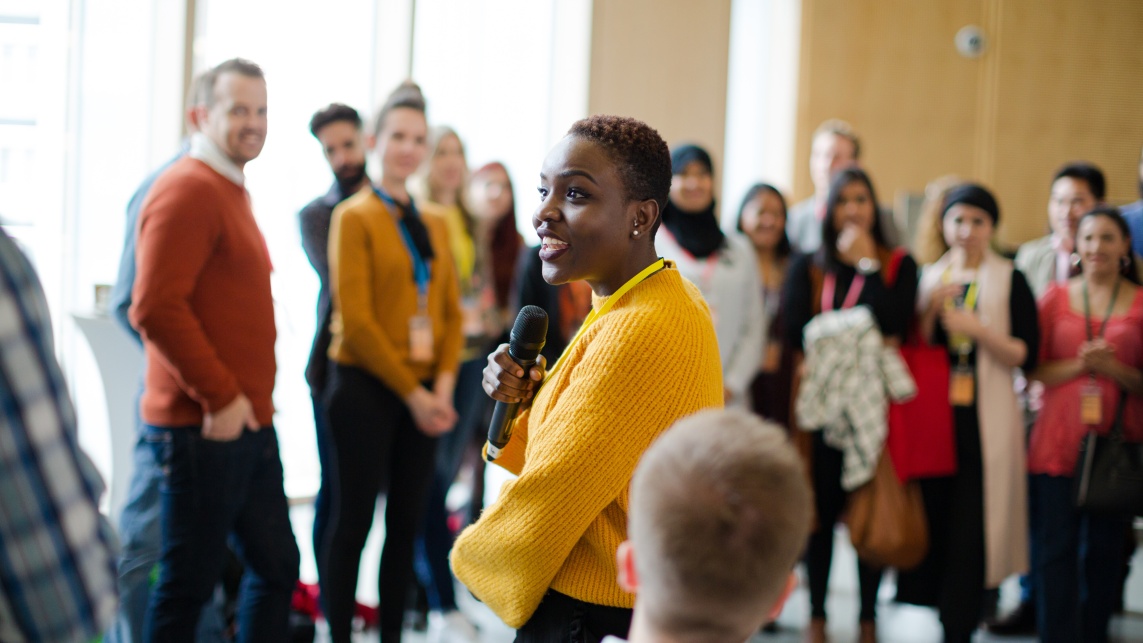 Woman holding a microphone speaking at an event to a group of people