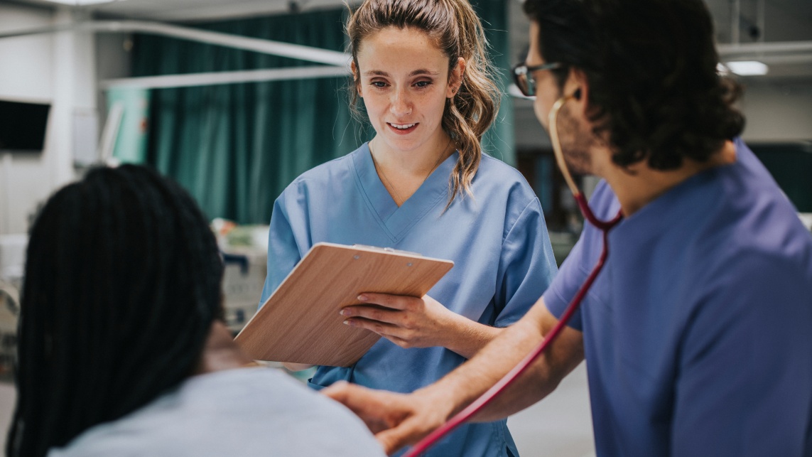 A male doctor examining a patient with a stethoscope. The female doctor writes notes on a clipboard. 