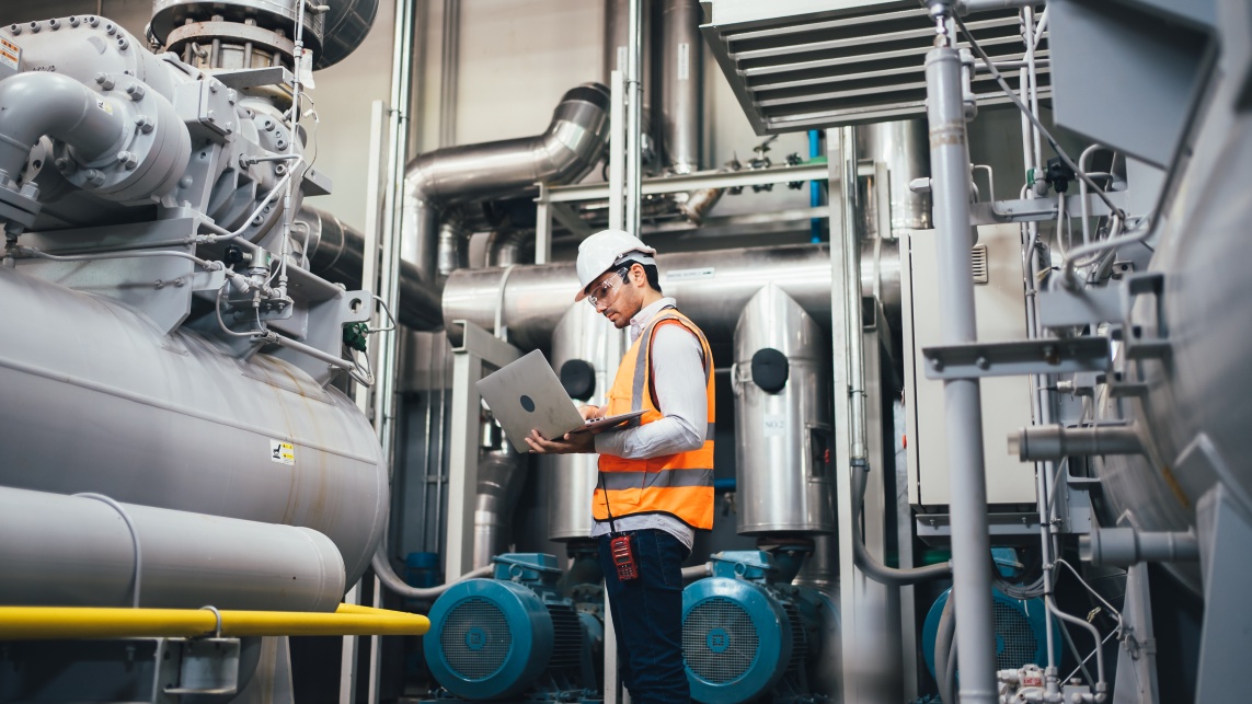 A man wearing high visibility clothing and a hard hat is holding a laptop and is surrounded by machinery and pipes.