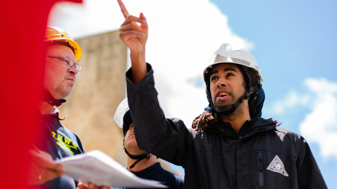 Male apprenticeship student is on a construction site pointing his index finger to the left. Next to him, a male holds papers in his hands and wears a yellow safety hat.