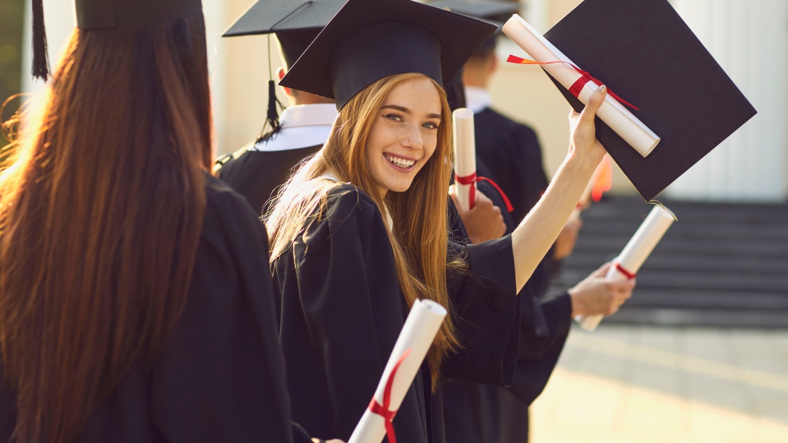 A group of graduates, one is a woman looking back and smiling whilst holding a certificate and scroll in the air.