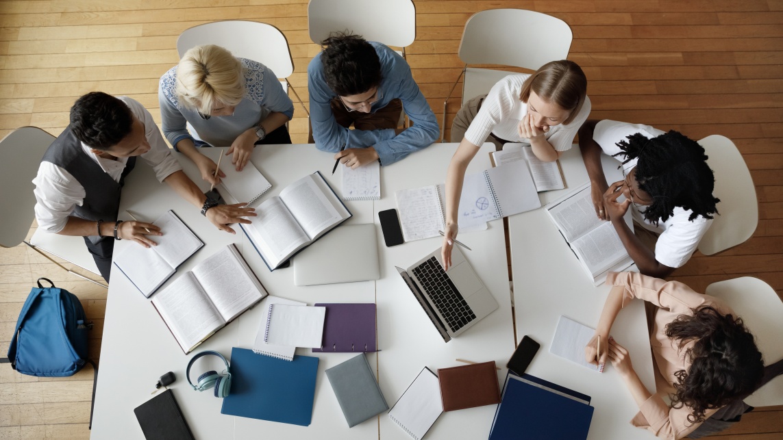 A birds eye view of desk with six students working together with their books and laptops spread across the table. 