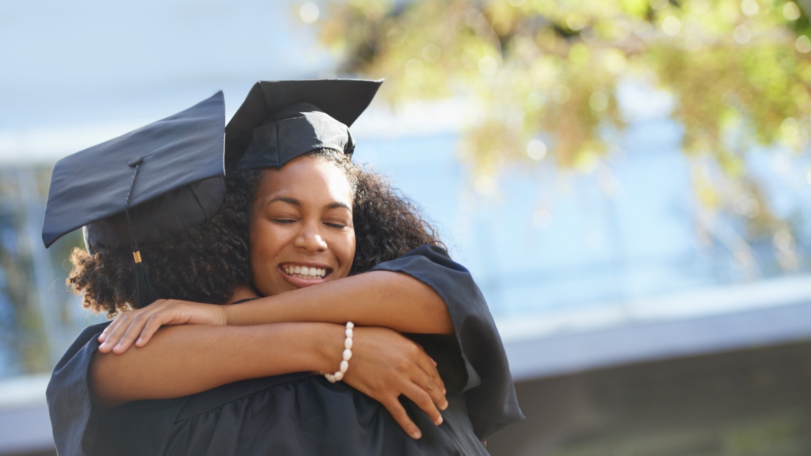 Two graduates smiling and hugging outdoors. 
