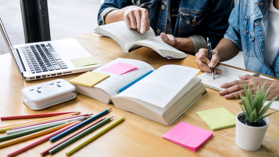  A desk with colorful stationary, a book, laptop, potted plant and two people working together from a book and notepad. 