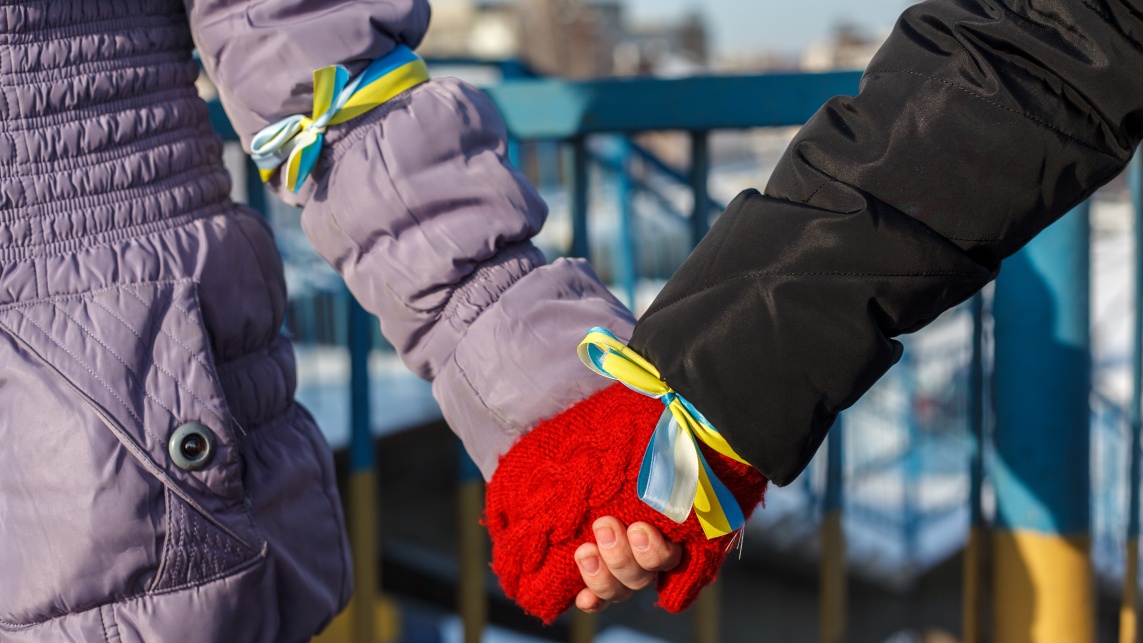 Two people wearing coats and gloves holding hands. Ribbons tied around their arms are in the blue and yellow colours of Ukraine's flag.