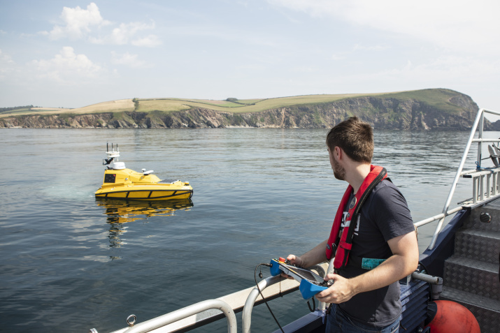 Man wearing a life jacket uses a handheld controller while looking at a small unmanned vessel at sea