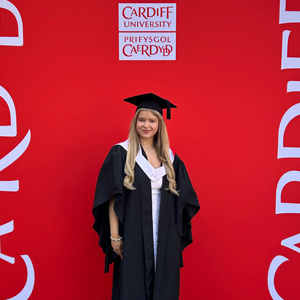 Lolli Lifting founder stands against a Cardiff University red backdrop. She wears a graduation gown and cap.