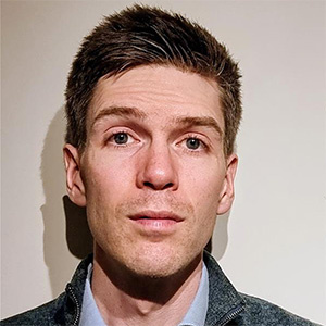 Charlie Ogilvie stands in front of a white wall. Charlie wears a grey sweater and blue shirt.
