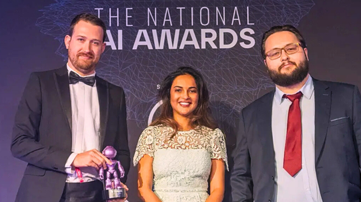 Hertzian founders stand in front of a National AI Awards banner. The founder on the left is holding an award statue.