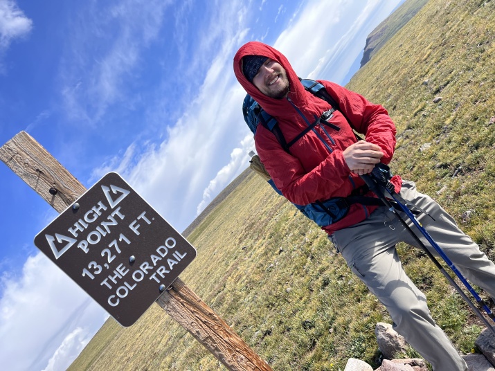 Darren Tindall at the highest point of the Colorado Trail standing next to a sign stating that wearing a red coat