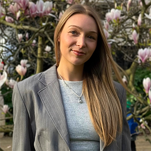 Darcy stands outside in front of a flowering tree. Darcy wears a grey top and blazer. 