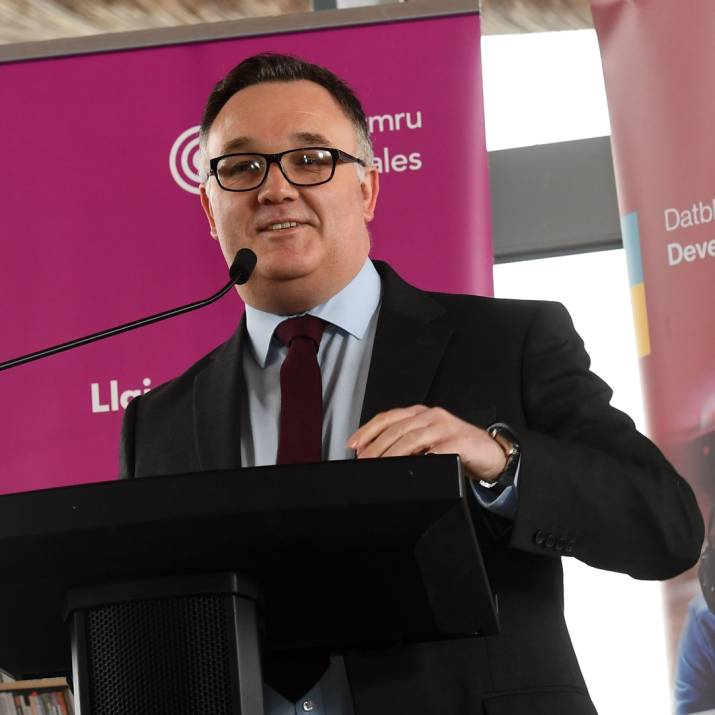 David Hagendyk, CEO of Colegau Cymru (Colleges Wales) stands in front of a pink pull-up banner and behind a lecturn and microphone. David wears a navy suit and glasses.