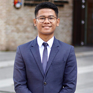 Dimas AlMaruf stands in front of a university building. Dimas wears a navy suit, white shirt and navy tie.