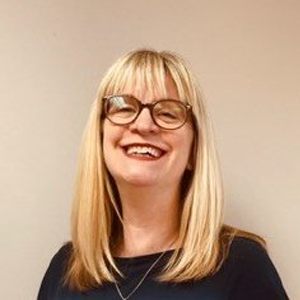 Emily Bird smiling at the camera against a cream background. Emily wears a black top and glasses.