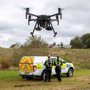 Two police officers stand in front of a police vehicle, operating a drone that is in the air.