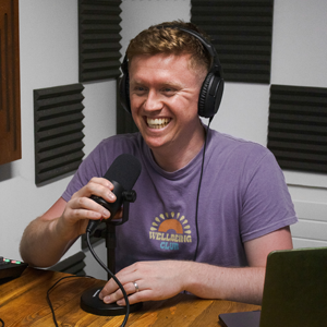 Founder of Engineer a Career, Josh Robertson sits in a podcast studio in front of a black and white checkered wall. Josh wears a purple top and is holding a microphone.