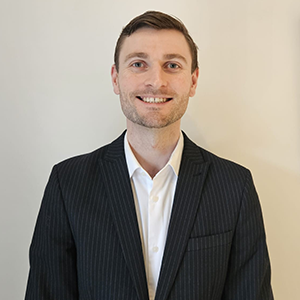 Leo Johnson stands in front of a white wall. Leo weares a black suit jacket and white shirt.