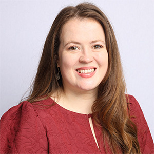 Lily Turvey stands in front of a pale background. Lily wears a red top and is smiling at the camera.