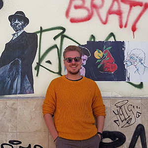 Photo of Oscar standing against a wall with grafitti, wearing sunglasses and an orange jumper