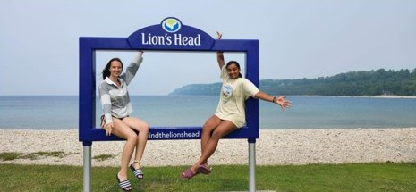 Two women pose, arms spread wide and sitting on a frame showing the text 'Lion's Head', with a beach and sea in the background.