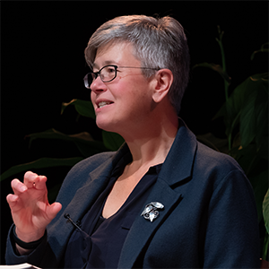 Professor Sasha Roseneil making a speech. She wears a navy blazer and stands in front of a black backdrop.